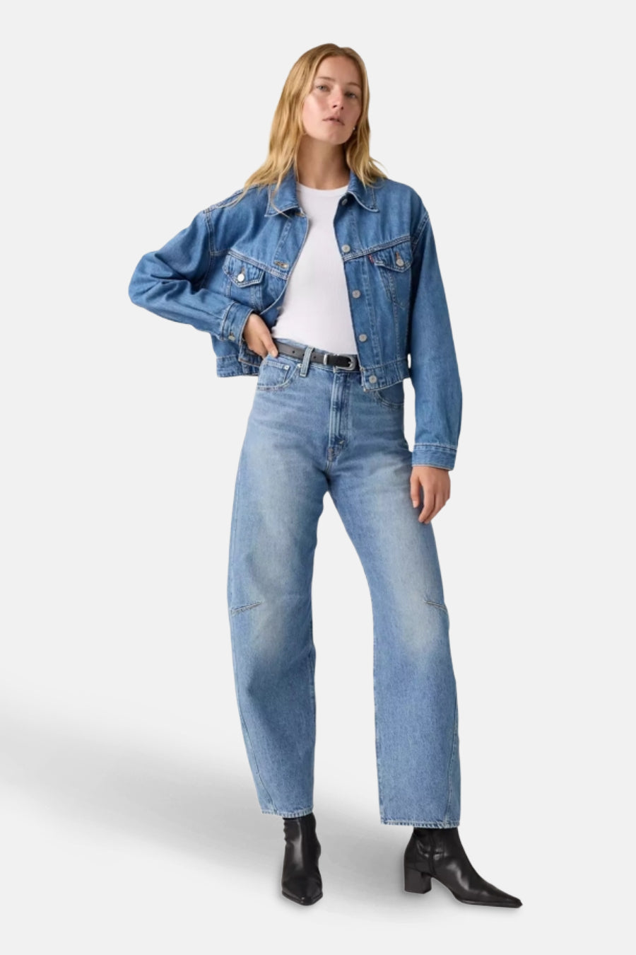 Vrouw in denim jack, witte top, blauwe jeans met riem en zwarte laarzen.