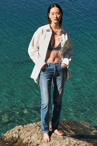 Lichtblauwe denim straight jeans van MAC, gedragen met een open linnen overhemd en een denim top op een rots bij het water.
