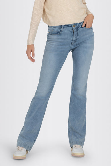 Bootcut jeans van lichtblauw denim van MAC, met een hand in de zak, gecombineerd met een beige trui en witte sneakers.
