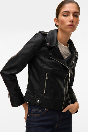 Manteau de cuir noir VERO MODA® avec des fermetures éclair argentées, porté sur une blouse blanche à bordures et des jeans noirs.
