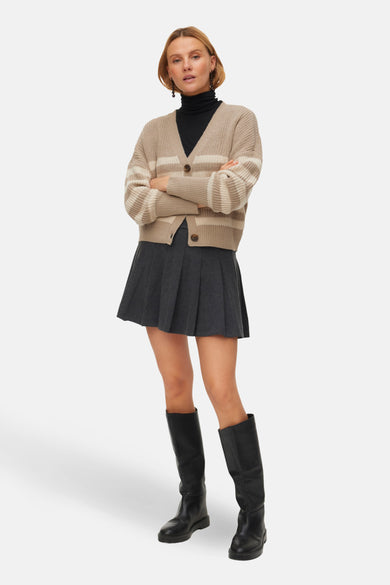 Beige cardigan van VERO MODA, met crèmekleurige strepen, gedragen over een zwarte coltrui en een grijze plooirok met zwarte laarzen.
