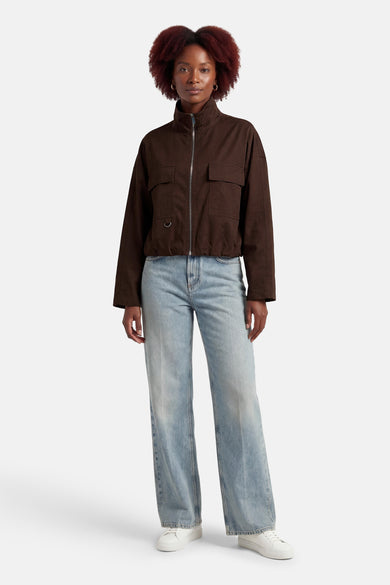 Bruine jas van OPUS, met lange mouwen en zakken, gecombineerd met een blauwe jeans en witte sneakers.
