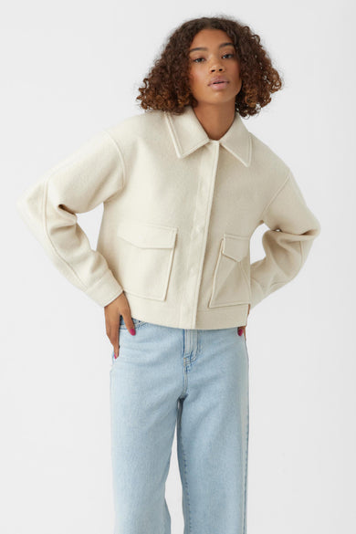 Beige korte jas van VERO MODA® met dubbele borstzakken, knoopsluiting en opstaande kraag, gedragen met lichtblauwe jeans.