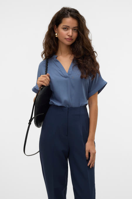 Blauw blouse met korte mouwen van VERO MODA, met V-hals en gedragen met donkerblauwe pantalon en zwarte tas.
