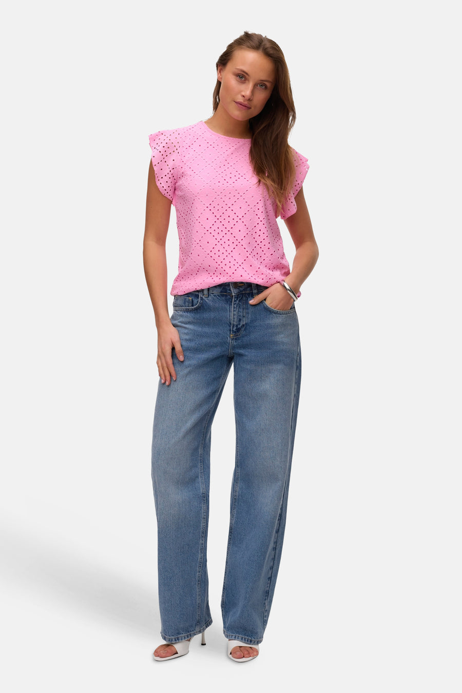 T-shirt rose à manches courtes de VERO MODA®, avec un motif ajouré et combiné avec un jean bleu et des sandales blanches.