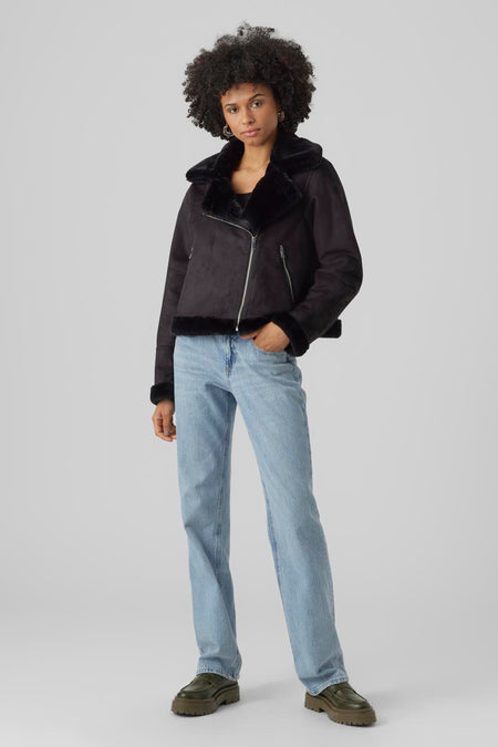 Zwarte korte jas van VERO MODA®, met een asymmetrische ritssluiting en een teddy kraag, gecombineerd met een blauwe jeans en groene schoenen.
