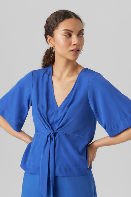 Blauwe blouse van VERO MODA, met knoopdetail en korte mouwen, gecombineerd met een blauwe broek.
