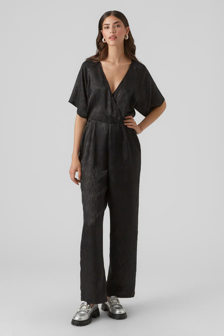 Zwarte jumpsuit van VERO MODA, met korte mouwen en een wikkelmodel, gecombineerd met zilverkleurige schoenen met decoratieve gespen.
