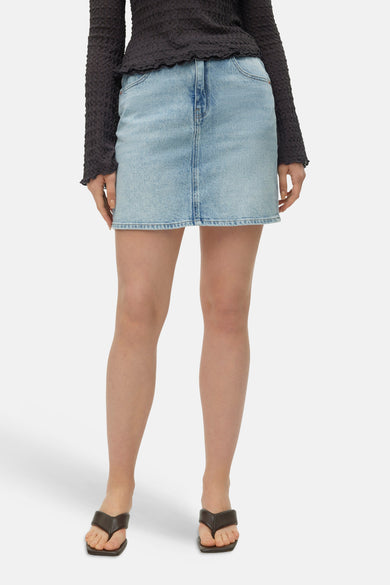 Lichtblauwe spijkerrok van VERO MODA, gedragen door een model met een donker getextureerd shirt en sandalen.
