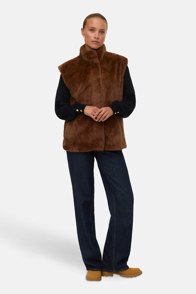 Bruine bodywarmer van VERO MODA, gedragen over een zwarte trui met gouden knopen en een donkerblauwe jeans.

