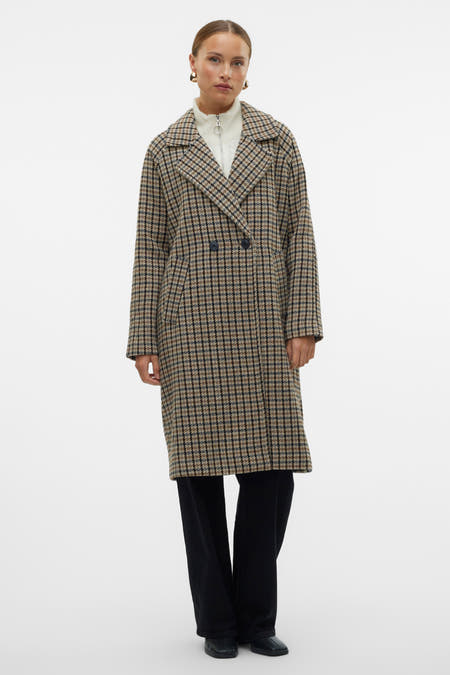 Bruine lange jas van VERO MODA, met een ruitpatroon en gedragen over een witte trui en zwarte broek.
