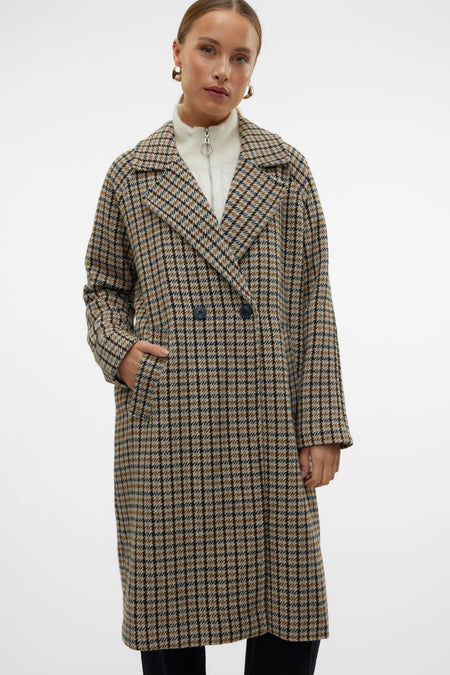 Bruine lange jas van VERO MODA, met een ruitpatroon, gedragen over een witte top met ritssluiting.
