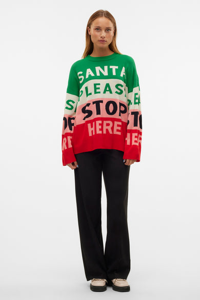 Multikleurige trui met ronde hals van VERO MODA, met de tekst "Santa please stop here" en gecombineerd met een zwarte broek en crèmekleurige schoenen.