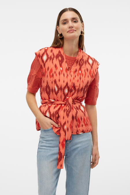 Oranje bodywarmer van VERO MODA® met ikatpatroon en strikceintuur, gedragen over een oranje top met openvallende kraag en jeans.
