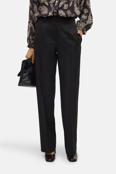 Pantalon noir de VERO MODA, avec des jambes droites, porté avec une blouse imprimée et un sac à main en cuir.