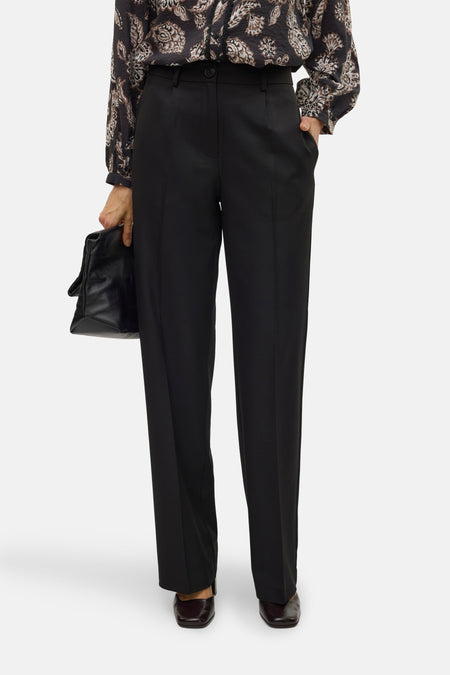 Zwarte broek van VERO MODA®, met rechte pijpen, gedragen met een bloes met print en een leren handtas.
