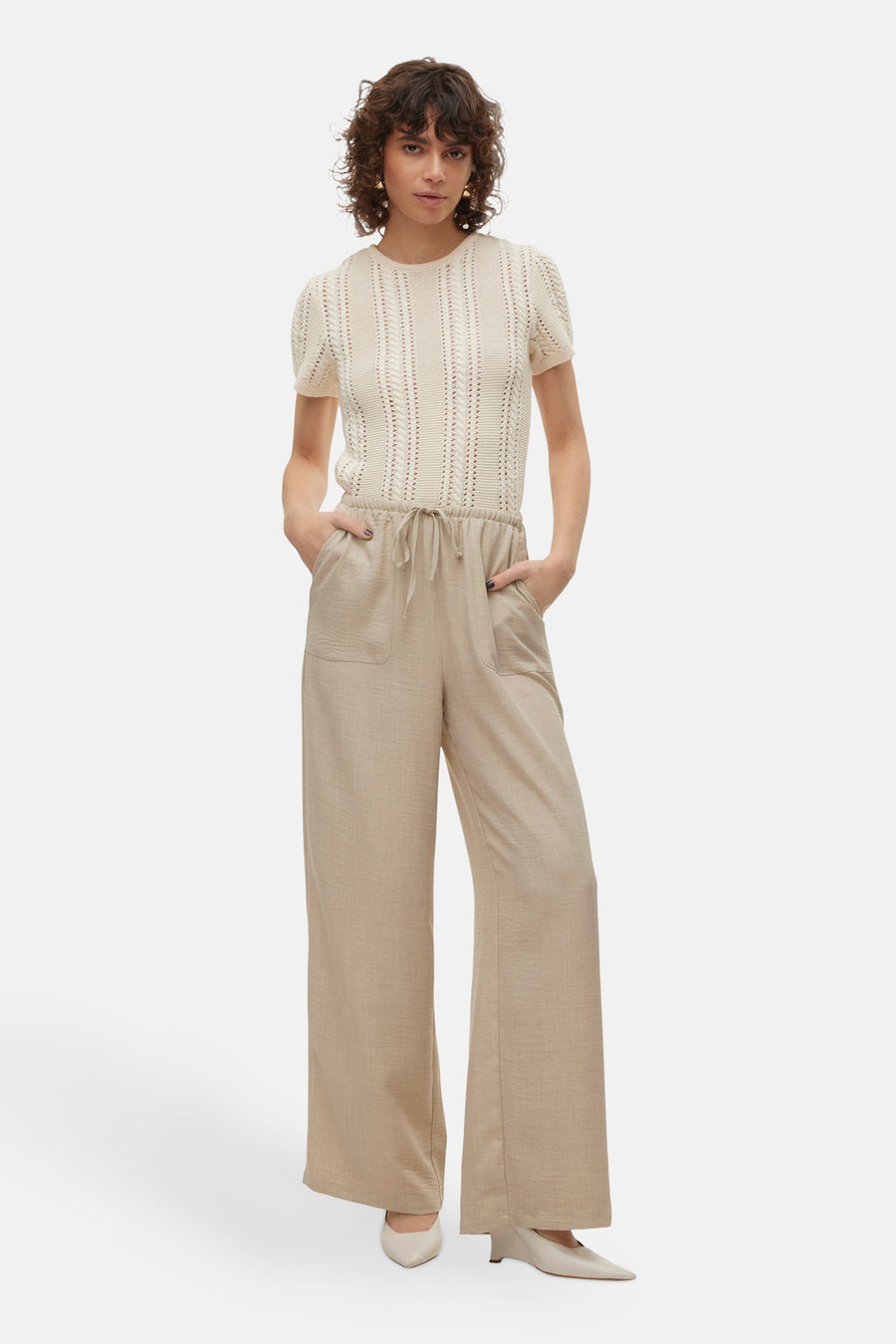 Jean beige de VERO MODA®, avec des jambes larges et des poches, porté avec un haut écru tricoté et des chaussures pointues.