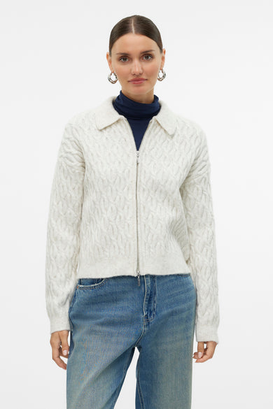 Ecrukleurig gilet van VERO MODA®, met kabelpatroon, gedragen over een donkerblauwe coltrui en een jeansbroek.
