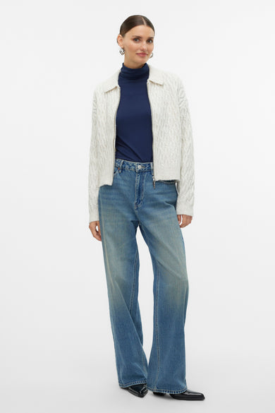 Ecru gilet van VERO MODA met kabelpatroon, gedragen over een donkerblauw coltrui en wijde jeans.
