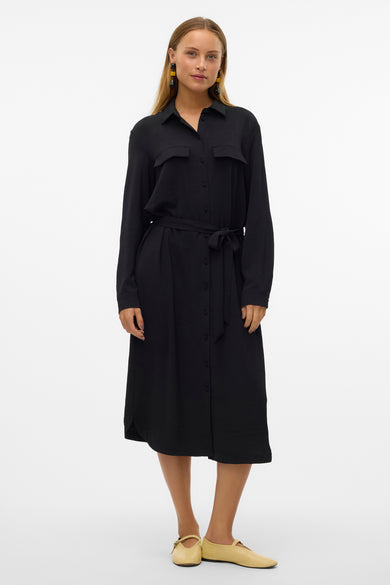 Zwarte jurk met lange mouwen van VERO MODA®, met knoopsluiting en riem in de taille, gedragen met platte schoenen.

