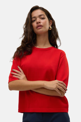 Pull rouge à col rond de Vero Moda, les bras croisés et boucles d'oreilles dorées.
