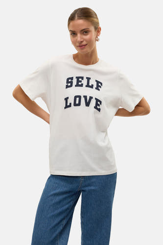Wit T-shirt met korte mouwen van VERO MODA, met donkerblauwe opdruk "SELF LOVE", gedragen met een blauwe jeans.
