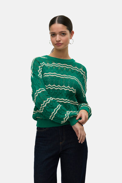 Groene trui met ronde hals van VERO MODA, afgewerkt met witte zigzagstrepen en lange mouwen, gecombineerd met donkere jeans en zilveren oorringen.

