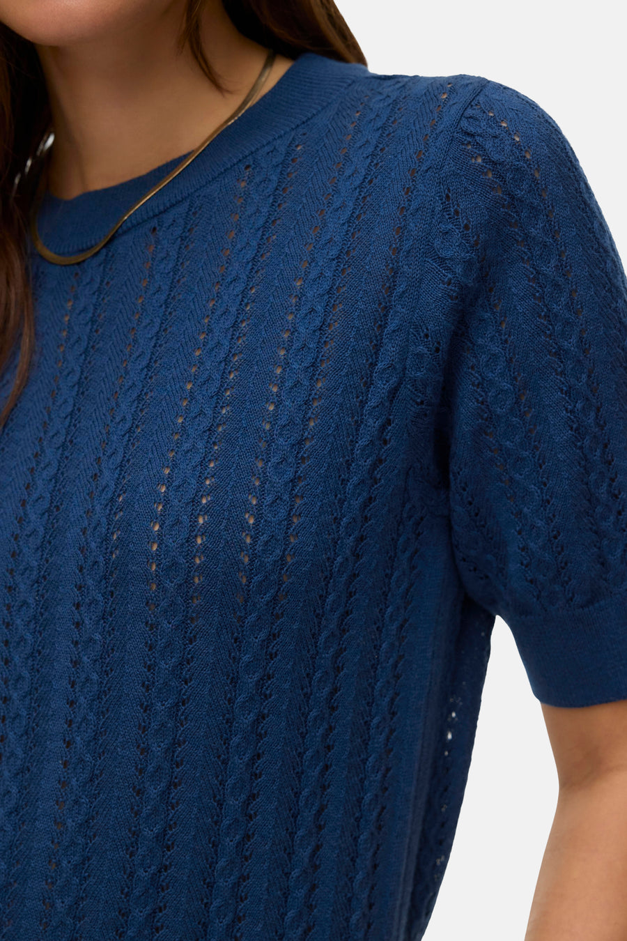 Pull bleu à col rond de VERO MODA®, avec motif ajouré et manches courtes, porté avec une chaîne dorée.