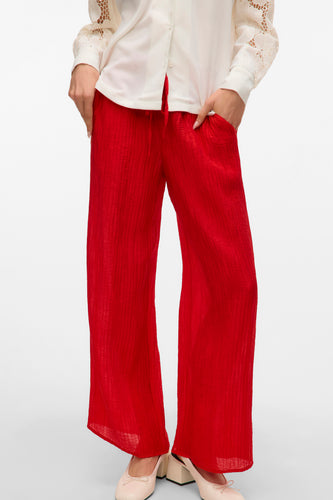 Rode broek van VERO MODA, met wijd uitlopende pijpen en textuur, gecombineerd met een witte blouse en platte schoenen.
