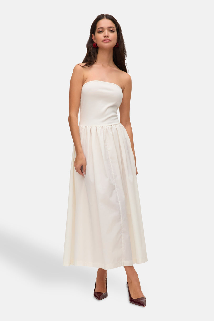 Vrouw in een strapless witte jurk met rode oorbellen en donkerrode hakken, staand op een witte achtergrond.