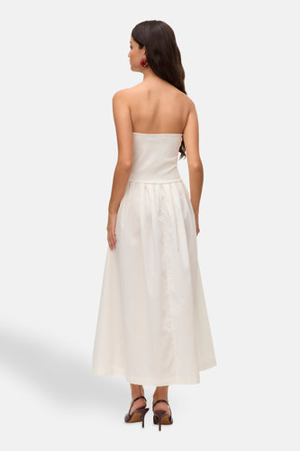 Achterkant van een model in een mouwloze witte jurk en bruine hakken tegen een witte achtergrond.