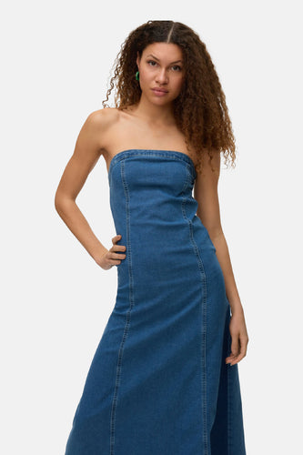 Vrouw in mouwloze denim jurk, staand voor witte achtergrond.