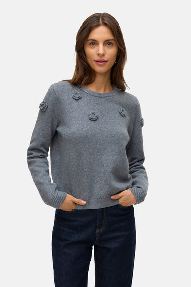 Grijze ronde hals trui van VERO MODA® met bloemendetails op de schouders en borst, gecombineerd met donkere jeans.
