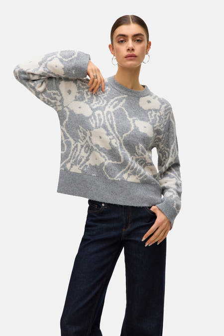 Grijze trui met ronde hals van VERO MODA® met bloemmotief, gecombineerd met donkere jeans en zilverkleurige oorbellen.
