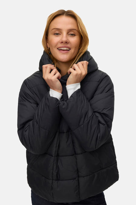 Zwarte korte jas van VERO MODA®, met lange mouwen en een capuchon, gedragen door een lachende vrouw.