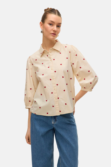 Chemise beige à manches courtes de Vero Moda, avec un motif de cœurs rouges sur toute la surface, associée à des jeans bleus.