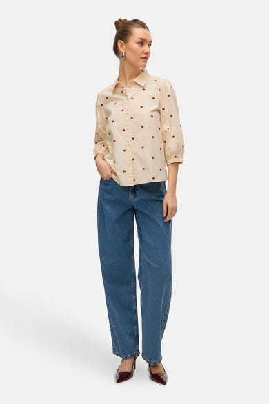 Blouse beige à manches courtes de Vero Moda, avec un imprimé de cœurs rouges, associée à des jeans larges.