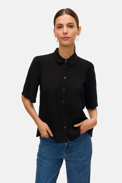 Chemise noire à manches courtes de VERO MODA, avec col boutonné et motif structuré subtil, portée avec des jeans.
