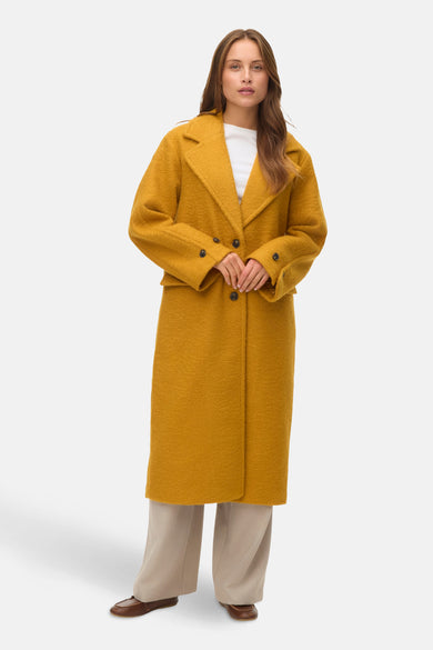 Gele jas van VERO MODA met lange pasvorm, gedragen over een wit shirt en beige broek.