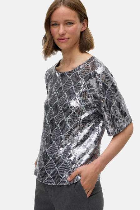 Blouse en argent avec des manches courtes de VERO MODA, ornée de paillettes et d'un motif à carreaux, portée avec un pantalon gris.
