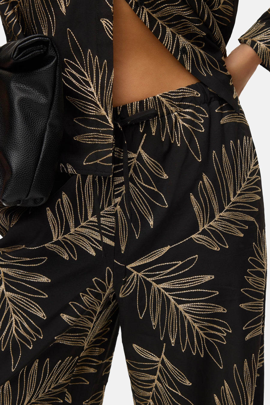 Close-up zwarte broek met gouden bladmotief, zwarte leren tas.