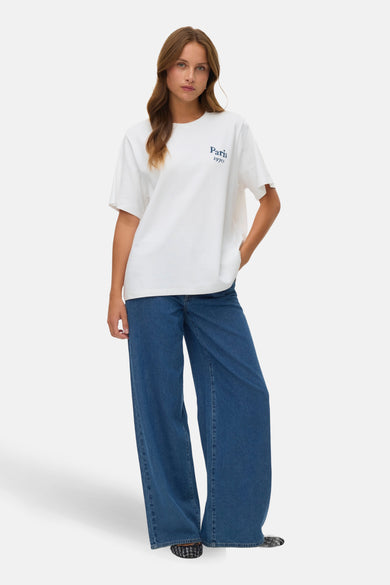 Wit T-shirt met korte mouwen van VERO MODA®, met 'Paris 1970' in blauw op de borst, gedragen met wijde jeans.
