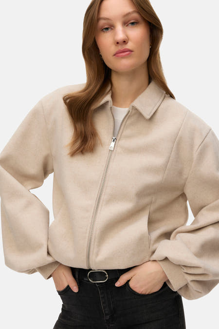 Ecru korte jas van VERO MODA, met zilverkleurige ritssluiting en steekzakken, gedragen met zwarte jeans en riem.
