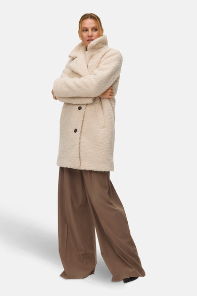 Beige lange jas van VERO MODA, met gekruiste armen gedragen over bruine, wijde broek.