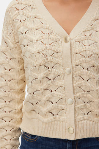 Gilet écru de VERO MODA avec fermeture à boutons et tricot ajouré, porté sur un jean bleu.