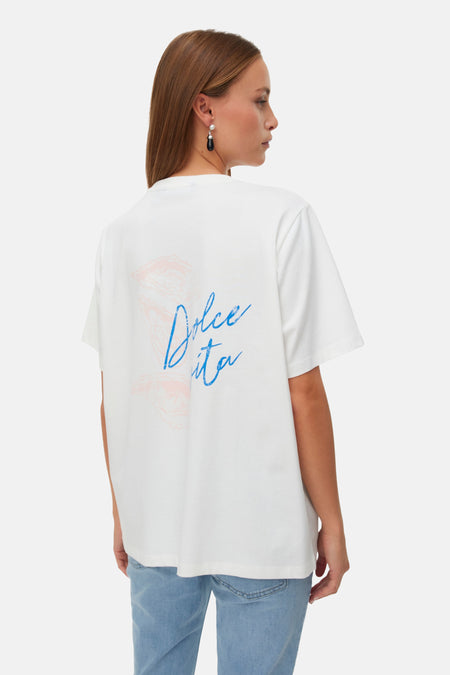 Wit T-shirt van VERO MODA, achterkant, met blauw 'Dolce Vita'-schrift en roze graphic, gedragen met lichtblauwe jeans.