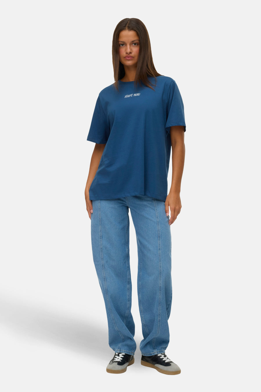 Vrouw, blauw T-shirt, spijkerbroek, staand.
