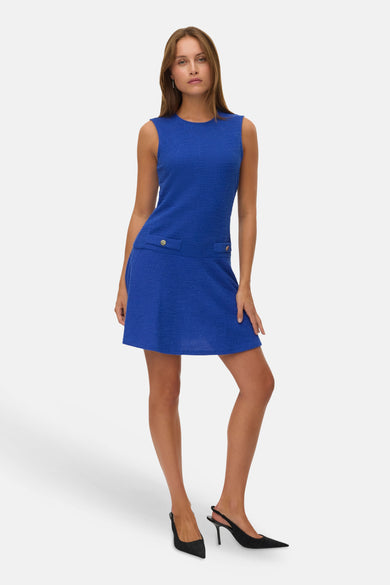 Blauwe mouwloze jurk van VERO MODA, met textuur en decoratieve knopen, gecombineerd met zwarte hakken.
