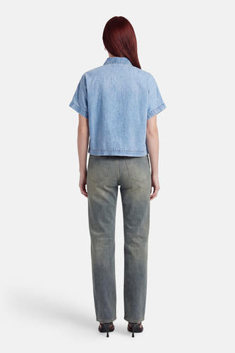 Chemisier en denim bleu clair à manches courtes de VERO MODA®, vu de dos sur un mannequin aux cheveux châtain roux, porté avec un jean gris foncé délavé et des sandales noires.
