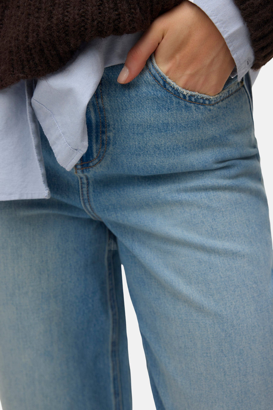 Lichtblauwe spijkerbroek van VERO MODA, close-up van een hand in de zak met gerafelde rand, gedragen met een bruine trui en blauw overhemd.